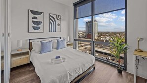 A modern bedroom with a large window, a bed with white linens, two bedside tables, and abstract wall art. - Boulevard Stays Modern Highrise STU Sky Pool, Gym 11 (Phoenix)