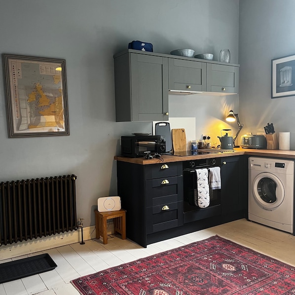 A compact kitchen with a washing machine, a small stool, and a red patterned rug.