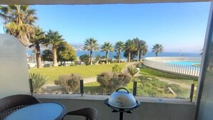 Un balcón con mobiliario de mimbre, una parrilla y vistas a una piscina y palmeras.