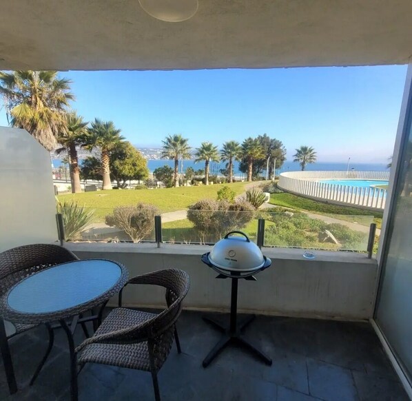 Un balcón con mobiliario de mimbre, una parrilla y vistas a una piscina y palmeras.