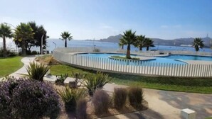 Una piscina con valla blanca, rodeada de follaje y palmeras, con vistas al mar y colinas al fondo.