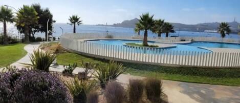 A pool with a white fence, surrounded by greenery and palm trees, with a view of the sea and hills in the background.