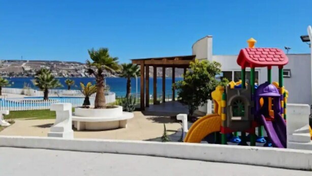 Un parque infantil con tobogán y estructuras para escalar, una zona de estar cubierta y vista al mar.