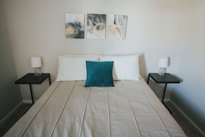 Una cama con un almohátero verde azulado, dos mesitas de noche con lámparas, y tres cuadros abstractos en la pared.