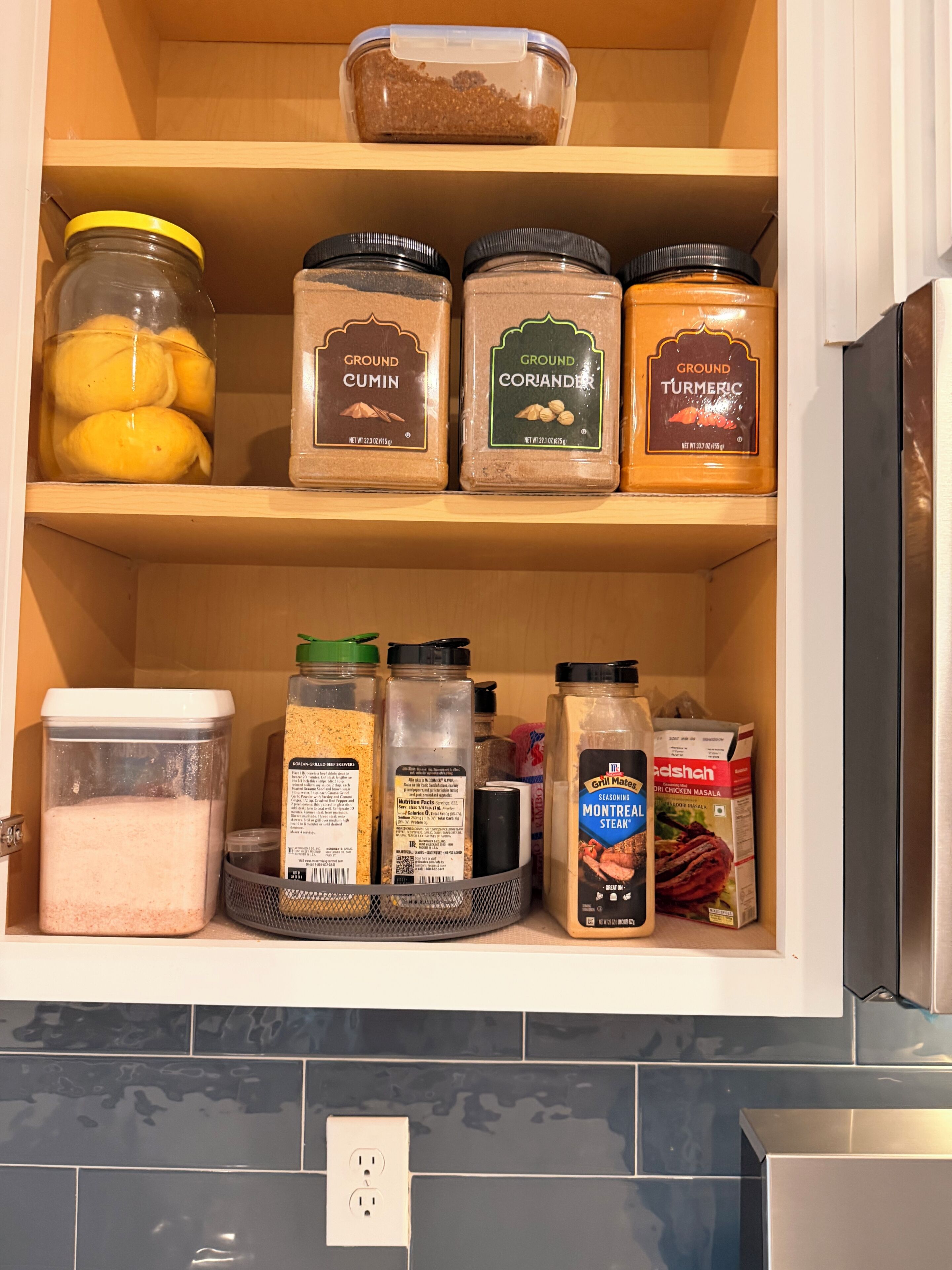 A kitchen pantry with various spices and condiments, including ground cumin, coriander, turmeric, and a container of lemon slices.