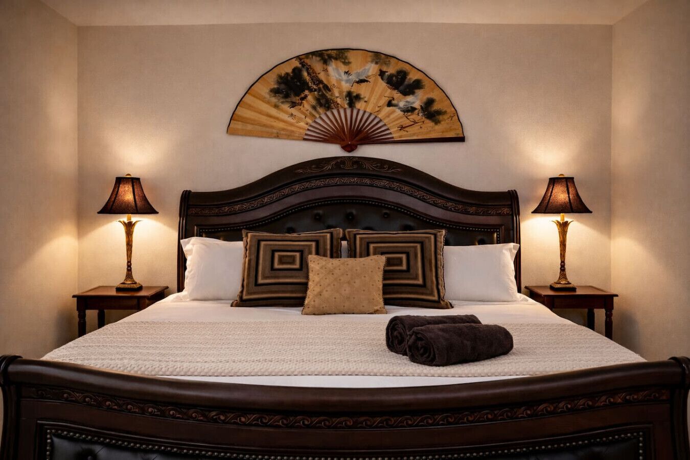 Uma cama com cabeceira de madeira escura, duas luminárias de apoio, um leque decorativo e um cobertor marrom.