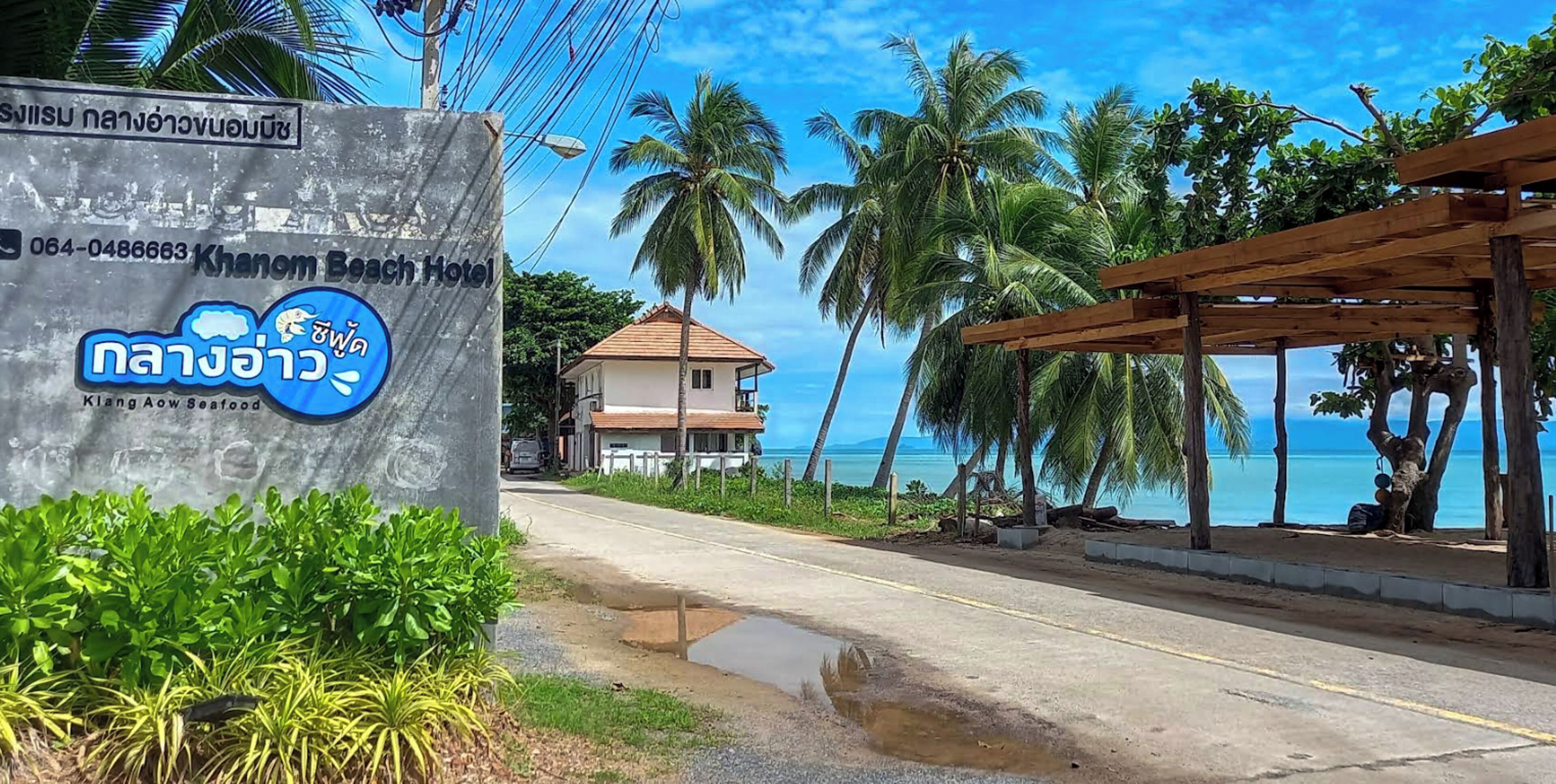 A beachfront hotel sign with a contact number, a view of the beach, palm trees, and a wooden structure.