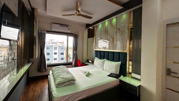 A hotel room with a bed, a ceiling fan, a window with curtains, and a view of buildings outside. - Hotel Runway (Barakpur)