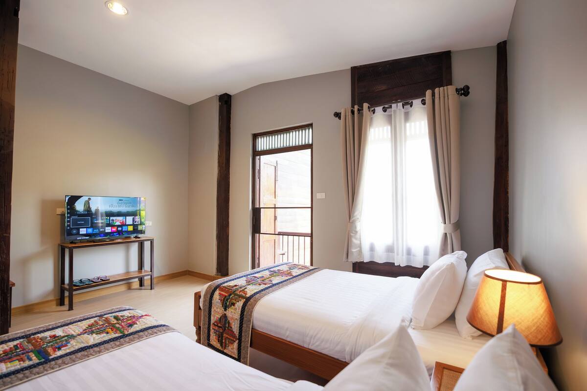 Une chambre d’hôtel avec deux lits, un téléviseur posé sur un meuble, une fenêtre avec des rideaux et une lampe sur la table de chevet.