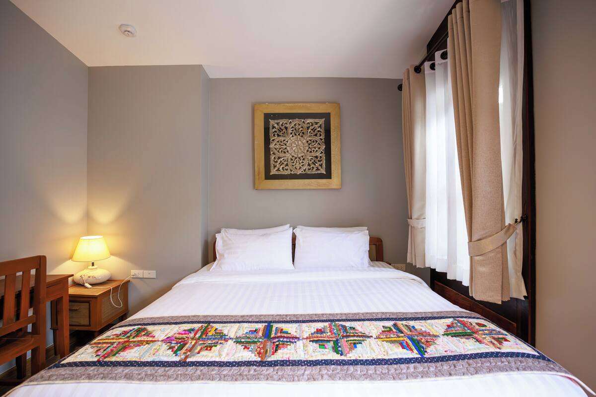 Un lit bien fait, avec une courtepointe, une lampe de chevet, une chaise et une œuvre d’art accrochée au mur.