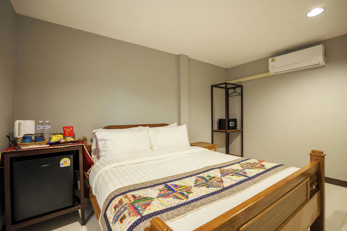 Un lit bien fait, recouvert d’une courtepointe, un petit réfrigérateur, une étagère avec quelques objets et un appareil de climatisation fixé au mur.