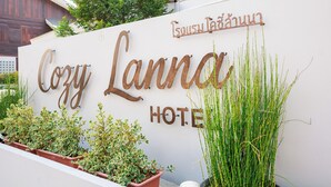 Une enseigne indiquant « Cozy Lanna Hotel », avec une inscription en thaï, entourée de plantes en pot et de verdure.