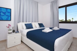 A modern hotel room with a large bed, white nightstands, a window with a view, and a blue abstract painting on the wall. - Greenery Villa - Large Private Pool (Protaras, Paralimni)