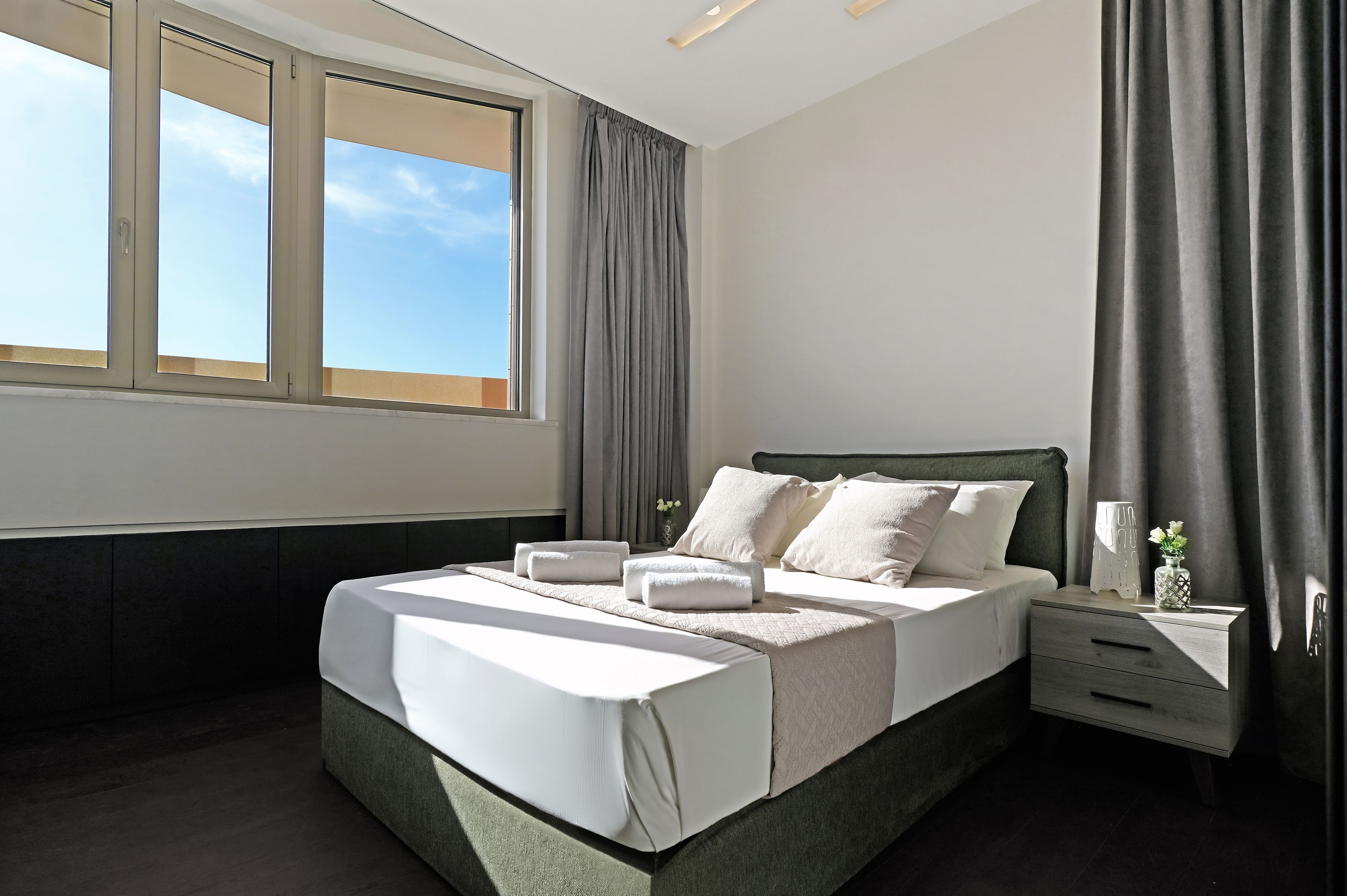 A modern hotel room with a large bed, a bedside table, a nightstand, and a window with curtains.