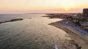 A coastal cityscape with a beach, a row of blue umbrellas, and a sunset. - Suncity Icon - Luxury Beachfront Villa (Ayia Napa)