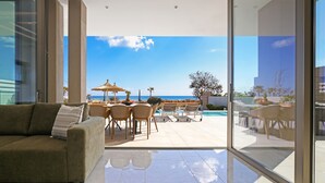 A modern living room with a glass door leading to a pool area, featuring a sofa, chairs, and a dining table. - Suncity Icon - Luxury Beachfront Villa (Ayia Napa)