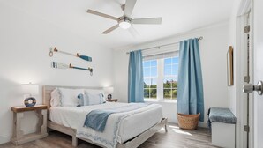Une chambre à coucher avec un lit, un ventilateur de plafond, une fenêtre avec des rideaux, une table de chevet et une porte.