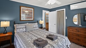 Een slaapkamer met een bed, nachtkastje, dressoir, plafondventilator, een ingelijst schilderij en een deur.