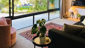 Un salon moderne doté de grandes fenêtres, d’un canapé, d’un fauteuil, d’une petite table avec une plante et d’un téléviseur posé sur un meuble.