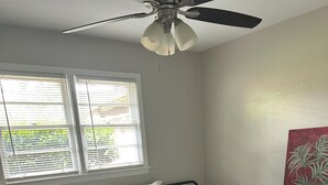 Ein Schlafzimmer mit Bett, Deckenventilator, Fenster mit Jalousien, Blumentopf und einem Bild an der Wand.