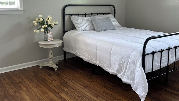 Ein Schlafzimmer mit einem Bett, einem Deckenventilator, einem kleinen Tisch mit einer Vase und einem Fenster mit Jalousien.