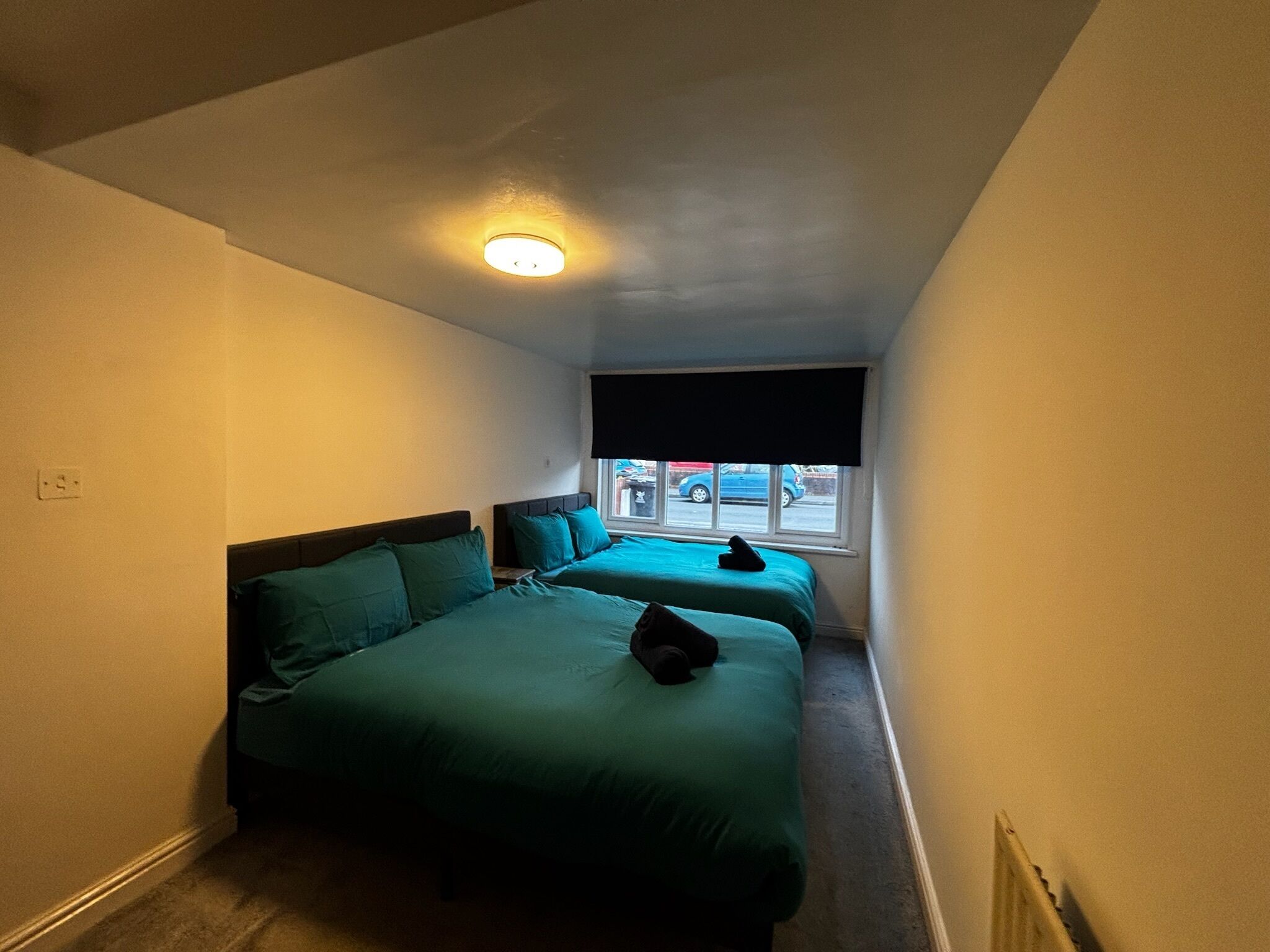 A small, narrow room with two beds, a window with a view of the street, and a ceiling light.
