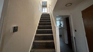 Una escalera que sube a un piso superior, con barandilla, un interruptor de luz en la pared y una puerta al final.