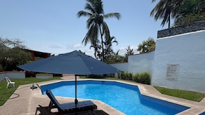 A swimming pool with a lounge chair and umbrella, surrounded by palm trees and a white wall. - Beautiful & Spacious 2 bed 2 bath Condo in Casa Bonita.  Ixtapa ,Zihuatanejo (Ixtapa Zihuatanejo)