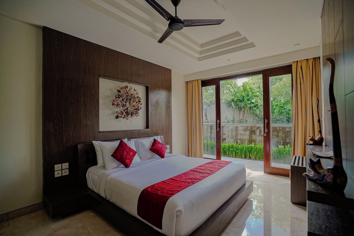 Une chambre d’hôtel moderne avec un grand lit, un ventilateur de plafond et une vue sur la verdure, visible à travers la porte vitrée.
