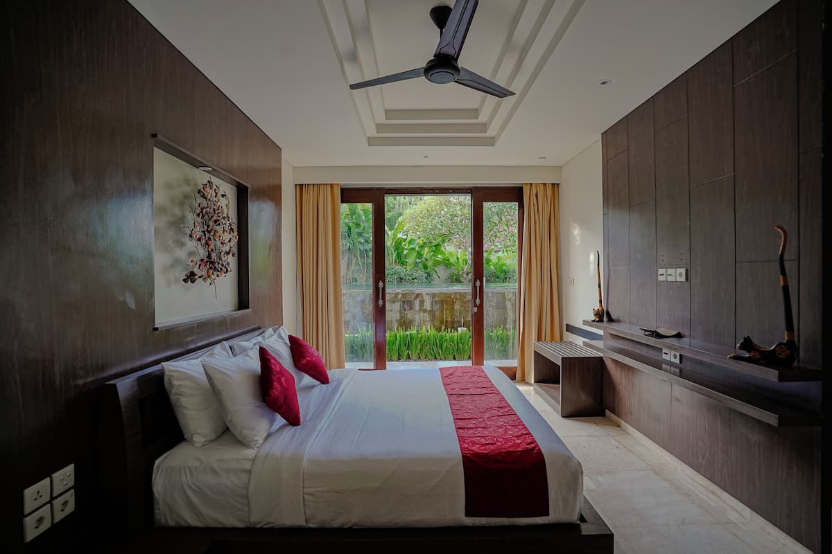 Une chambre à coucher avec un lit, un ventilateur de plafond, une fenêtre avec des rideaux et une vue sur la verdure.