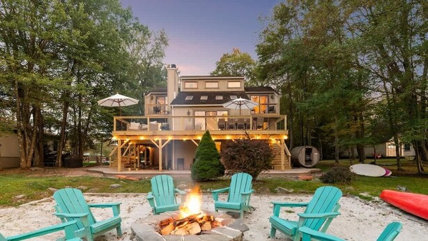 Ein zweistöckiges Haus mit einer Terrasse, einer Feuerstelle mit Sitzgelegenheiten und einem Kanu.
