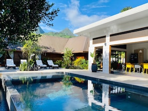 A modern pool area with lounge chairs, a dining area with yellow chairs, and a mountain view.