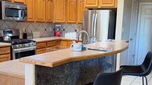 Cozinha moderna com armários de madeira, geladeira de aço inoxidável, micro-ondas e fogão a gás. Há dois banquetas pretas e uma ilha grande com bancada de granito.