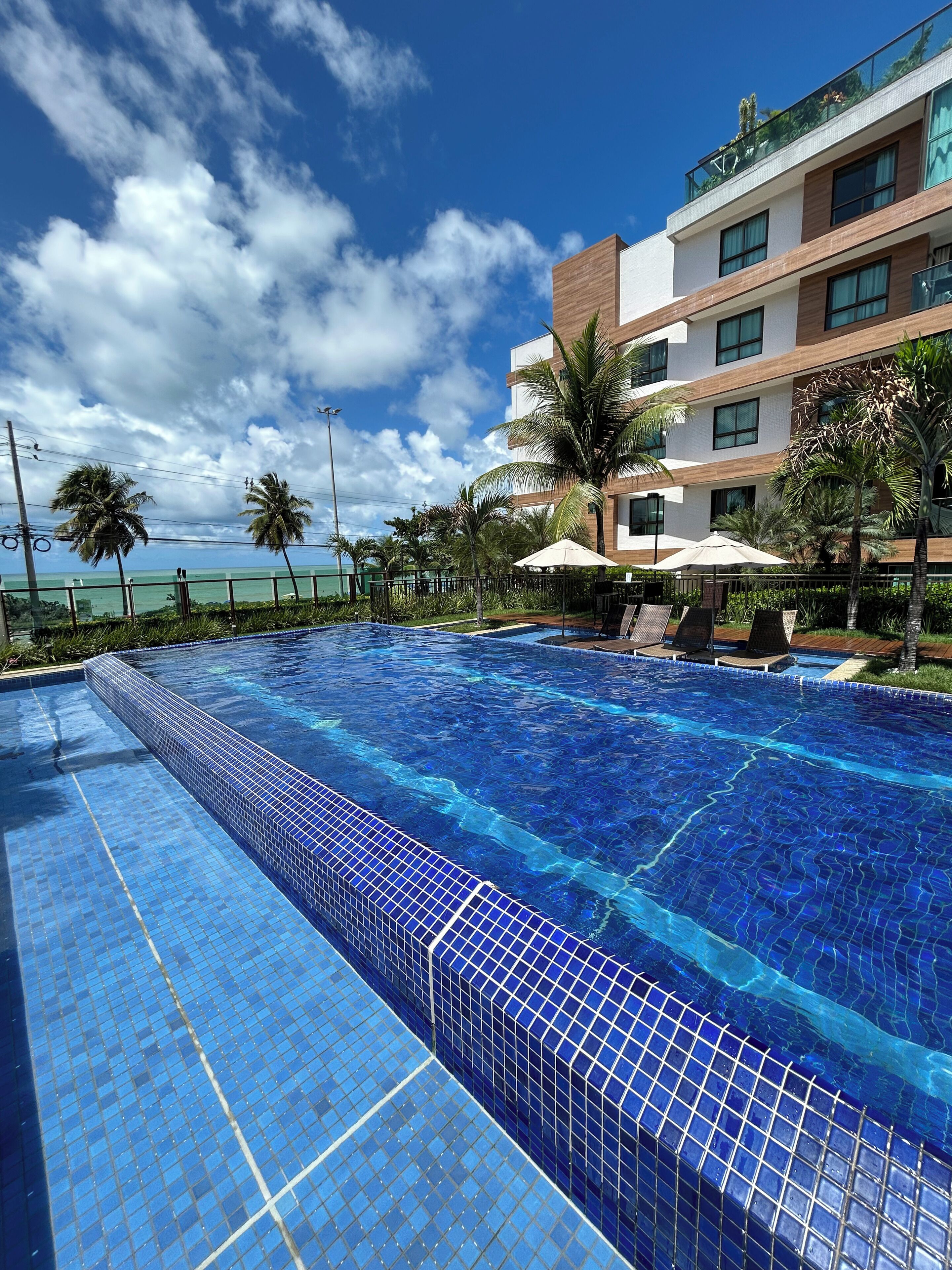Una piscina con piastrelle blu trasparenti, un edificio moderno con diversi piani e balconi, e palme.