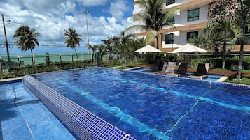 Una piscina con piastrelle blu trasparenti, un edificio moderno con diversi piani e balconi, e palme.