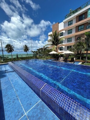 A swimming pool with clear blue tiles, a modern building with multiple floors and balconies, and palm trees. - Melhores Flats - Esplêndido beira-mar (João Pessoa)