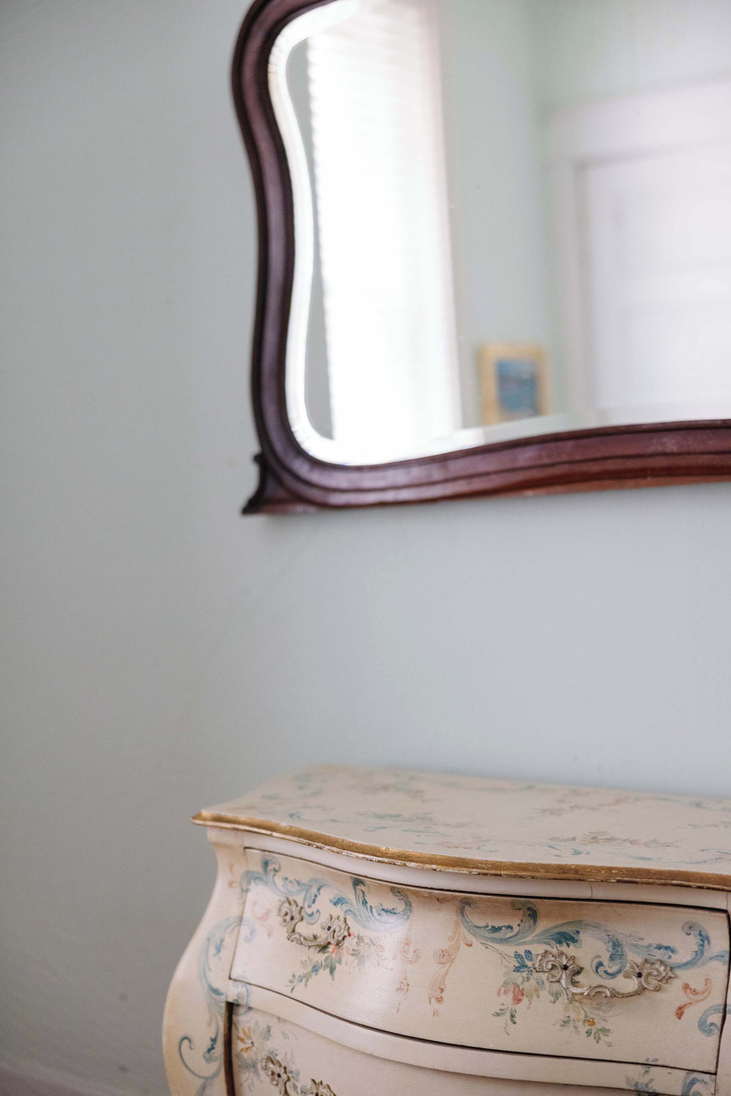 A vintage dresser with ornate floral patterns, a mirror with a dark frame, and a light-colored wall.