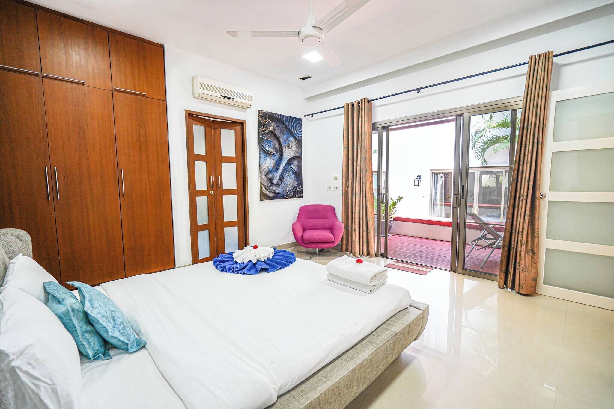 Habitación de hotel con cama, armario de madera, una silla rosa, un cuadro y una puerta corrediza de vidrio al exterior.