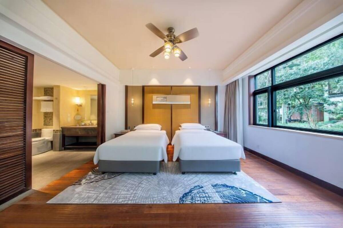 A hotel room with two beds, a ceiling fan, a large window, and a view of the outdoors.