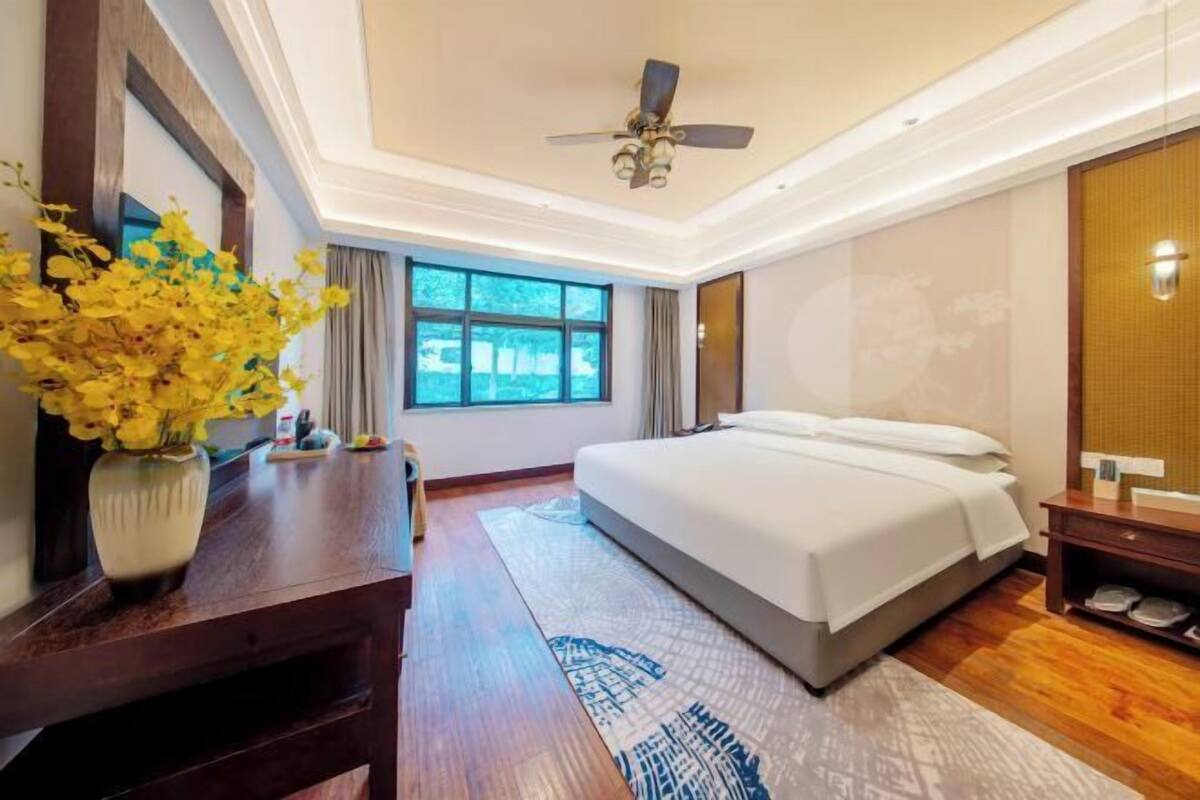 A hotel room with a bed, a desk, a vase with flowers, a ceiling fan, and a window.