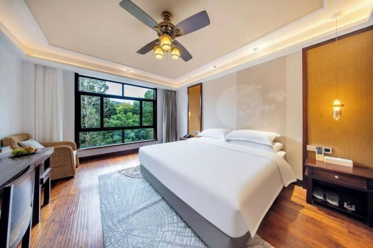 A hotel room with a large bed, a desk, a chair, a ceiling fan, and a large window with a view of greenery.