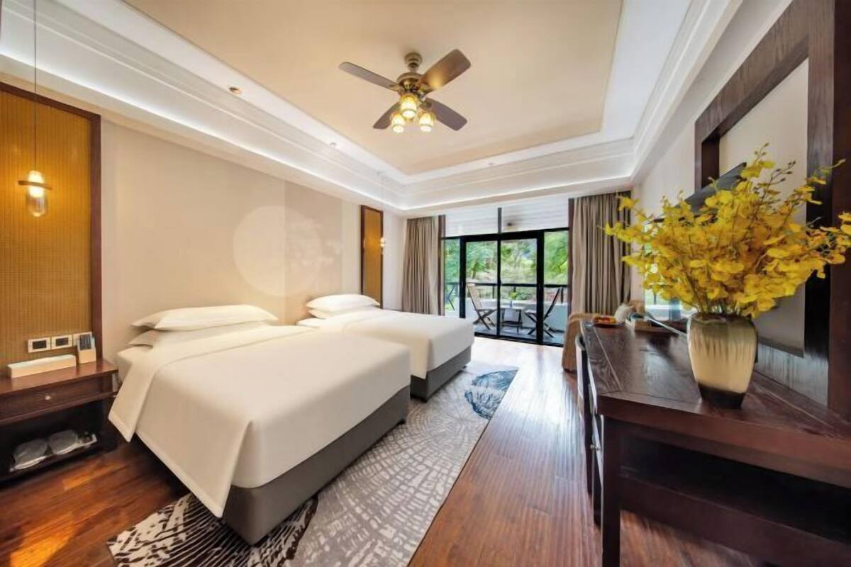 A hotel room with a bed, a desk, a chair, a ceiling fan, and a window with a view of greenery.