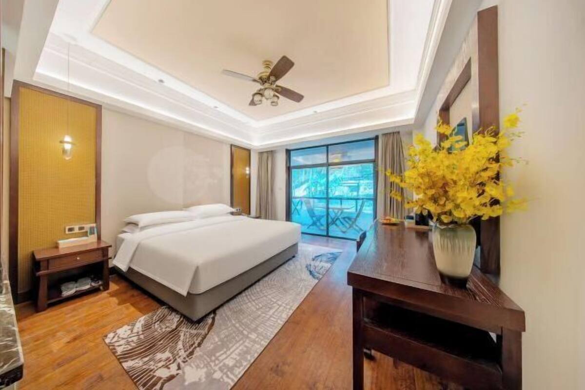 A hotel room with a bed, a desk, a chair, a ceiling fan, and a view of the outdoors.
