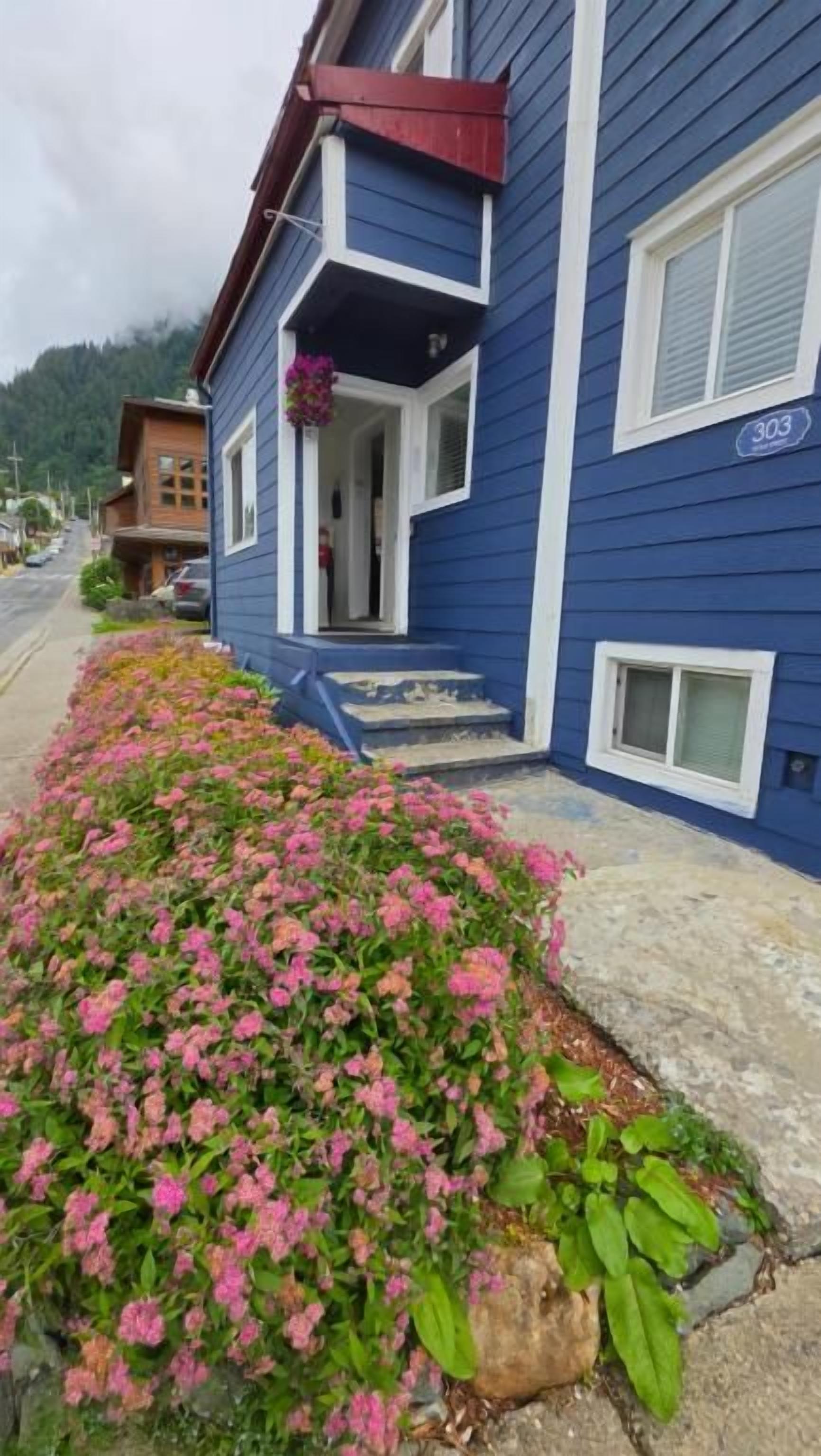Un edificio blu con il numero '303' sul lato, un portico con gradini e un'aiuola con fiori rosa.