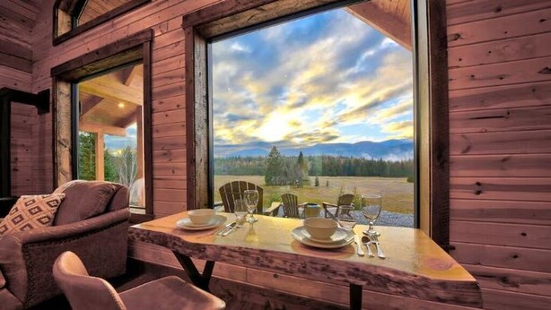 Een houten chaletinterieur met een eettafel gedekt voor twee personen, grote ramen en uitzicht op een bebost landschap.