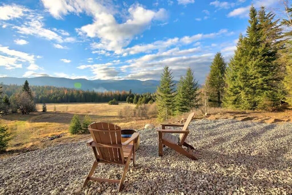 Two wooden Adirondack chairs on a gravel surface with a mountainous landscape and trees in the background.