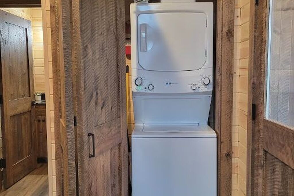 A laundry room with a stacked washer and dryer, wooden walls, and a door leading to another room.