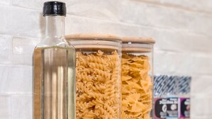 Drei Gläser mit Pasta auf einer reflektierenden Oberfläche vor einer weißen Ziegelwand.