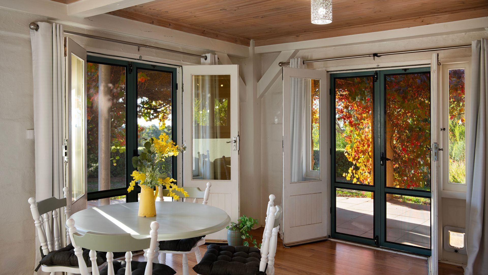 A room with a dining table, chairs, a vase with flowers, and a view of autumn foliage through large glass doors.