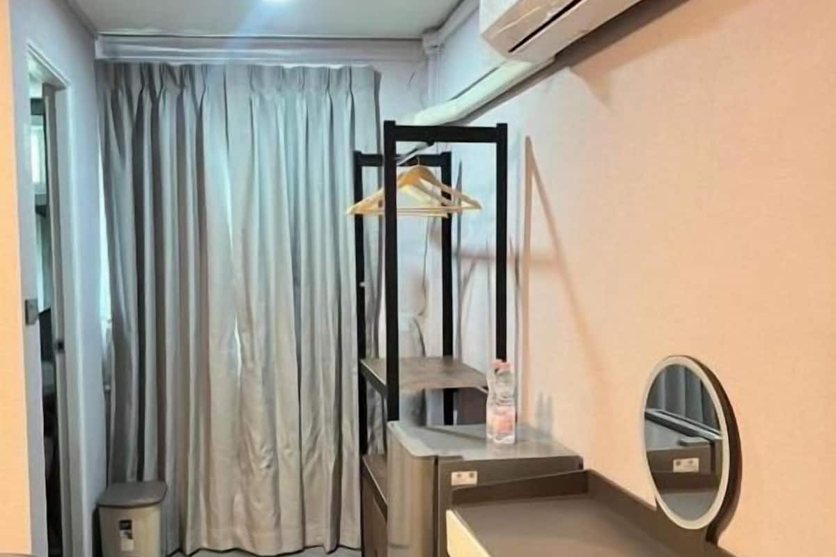 Quarto compacto com cama, escrivaninha, cadeira, espelho, ar condicionado e um pequeno banquinho.